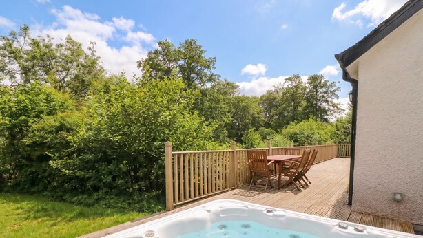 A hot tub on a wooden deck with a table and chairs, surrounded by greenery and a wooden fence. - Ghillie's Cottage (Killin)