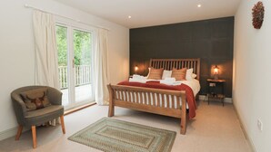 A bedroom with a bed, a chair, a rug, a nightstand with a lamp, and a view through a window. - Ghillie's Cottage (Killin)
