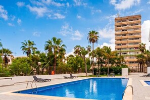 A swimming pool with lounge chairs, palm trees, and a tall building in the background. - Casa Miriam Apartment (Los Cristianos)