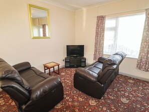 A living room with a sofa, a television, a small table, and a window with curtains. - Cottage 1 (Tenby)