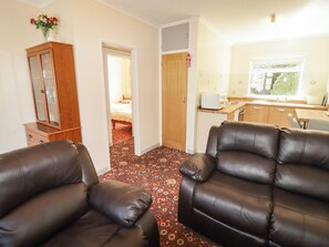A room with two leather sofas, a wooden cabinet, a kitchen area with a window, and a patterned carpet. - Cottage 1 (Tenby)