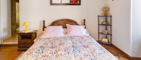 Un dormitorio con una cama de madera, una mesita de noche con una lámpara, una mesita con un marco de foto y una repisa de pared con un cuadro.