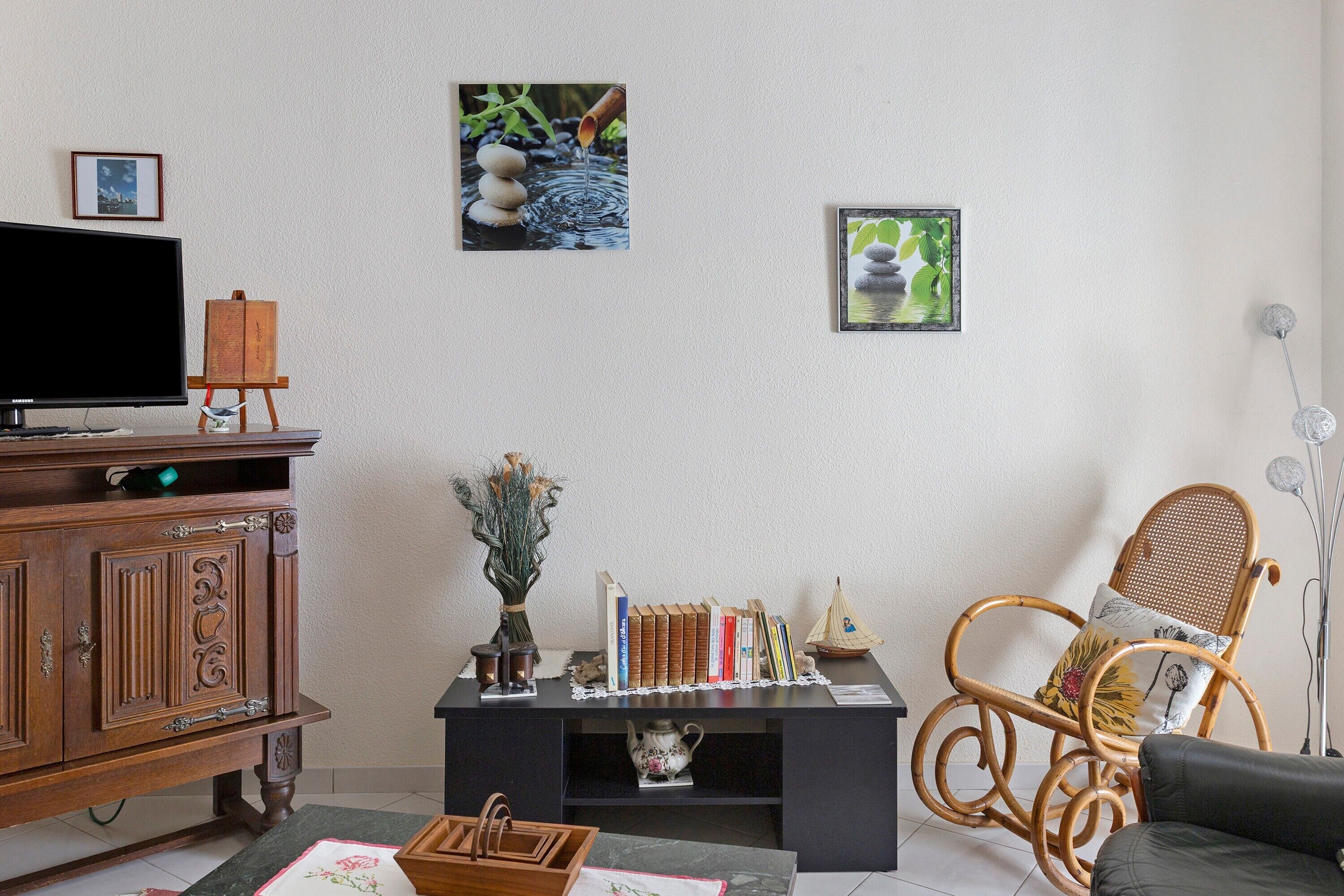 A living room with a television, wooden cabinet, books, a vase with dried flowers, a wicker chair, and wall art.