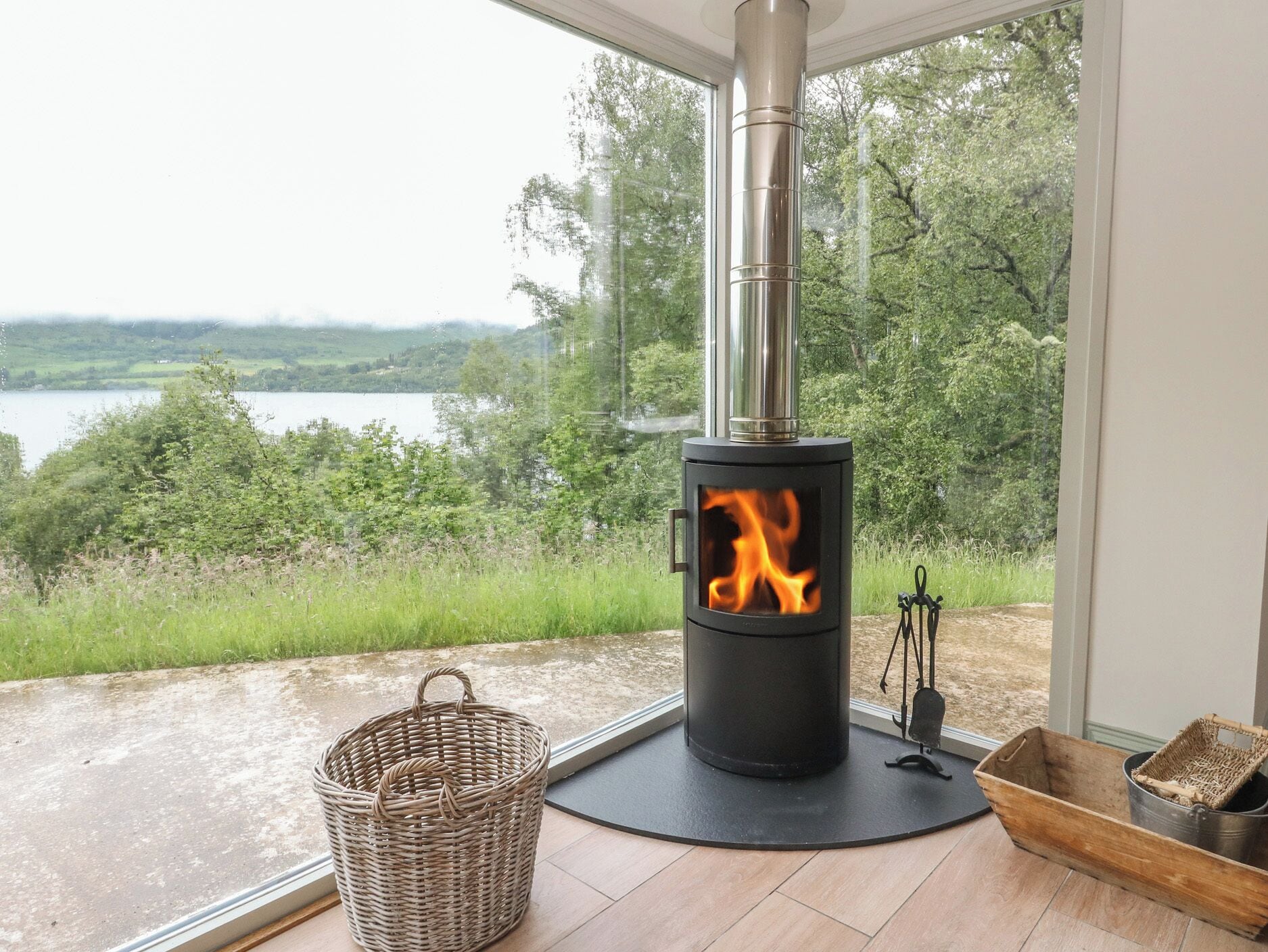 A wood-burning stove with a glass front, a wicker basket, and a metal container on a wooden floor by a window overlooking a lake.