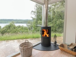 Una estufa de leña con frente de vidrio, una cesta de mimbre y un recipiente metálico sobre un piso de madera, cerca de una ventana con vistas a un lago.