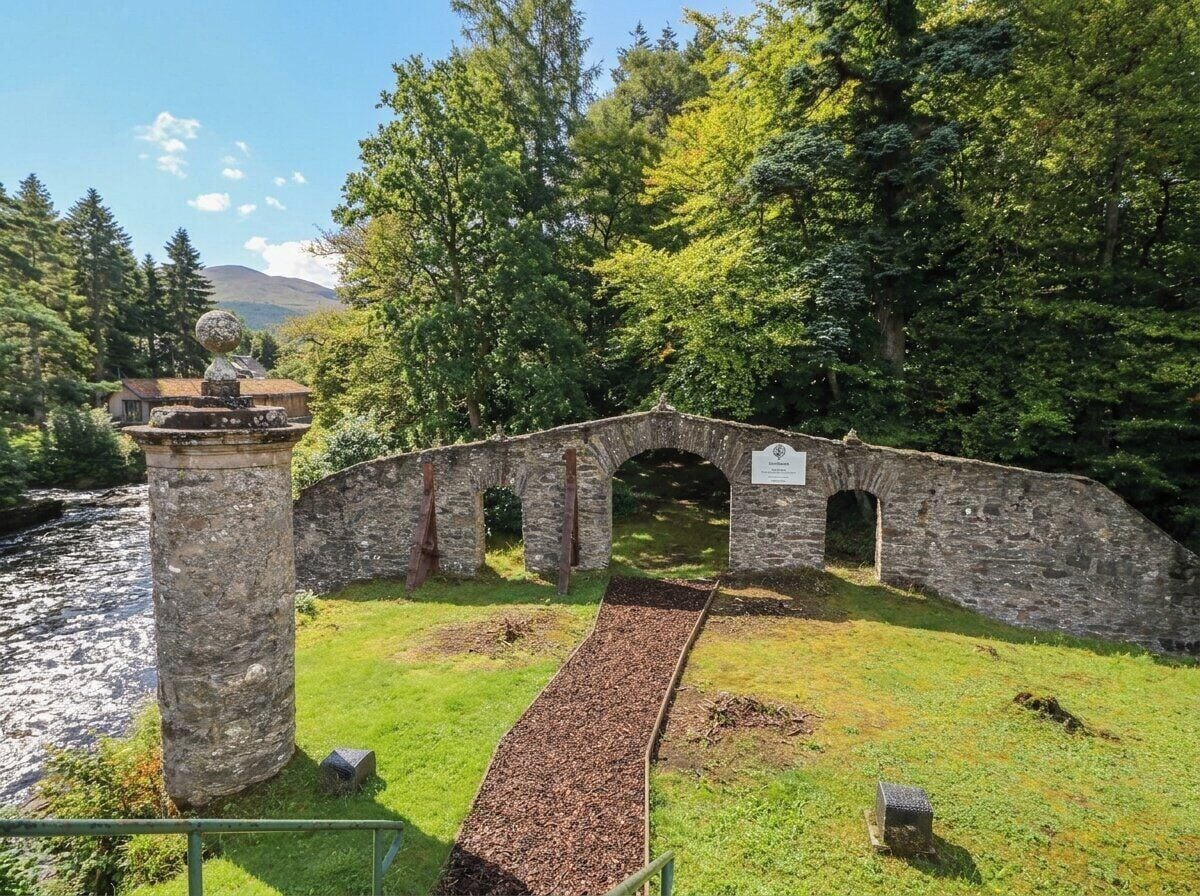 Un puente de piedra con arcos, una zona cubierta de césped y un río al fondo.