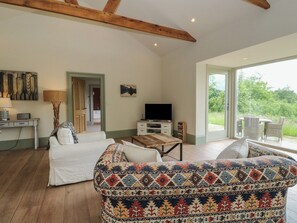 A spacious living room with a large window, a white sofa, a patterned ottoman, a television, and a dining area with wicker furniture. - The Tractor Shed (Aberfeldy)