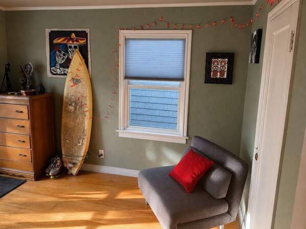 Una habitación con un aparador de madera, una tabla de surf, una ventana con persianas, un sillón gris con un cojín rojo y adornos en la pared.