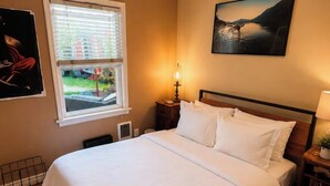 Ein Schlafzimmer mit einem großen Bett, einem Fenster mit Jalousien, einem Nachttisch mit Lampe und einem Gemälde an der Wand.