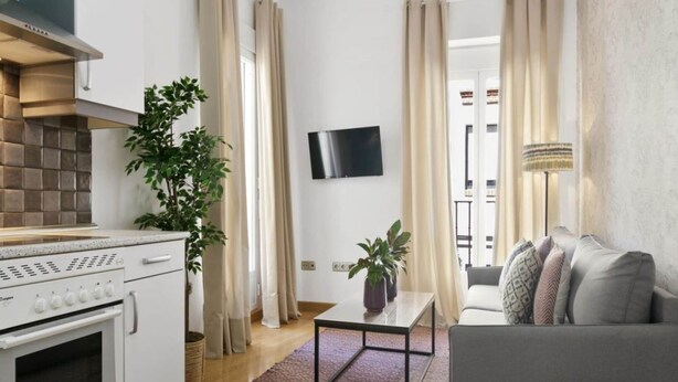 A modern kitchen with a stove, a dining area with a table and chairs, a grey sofa, and a TV mounted on the wall. - 8-guest Malasana Central Suite (Madrid)