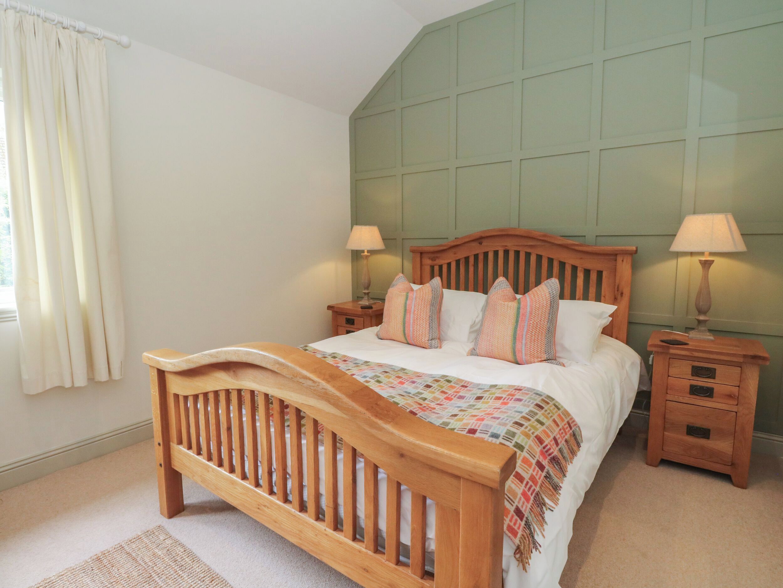 A bedroom with a wooden bed, two bedside tables with lamps, a window with curtains, and a green wall paneling.