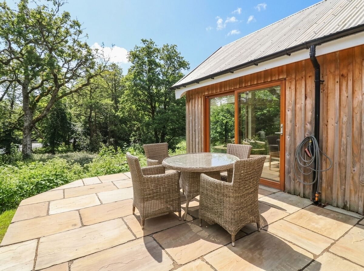 Eine Terrasse mit rundem Tisch und Korbstühlen, ein Holzhaus mit Glastür und ein Garten mit Bäumen und Sträuchern.