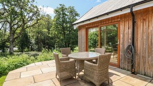 Une terrasse avec une table ronde et des chaises en osier, un bâtiment en bois avec une porte vitrée, et un jardin avec des arbres et des arbustes.