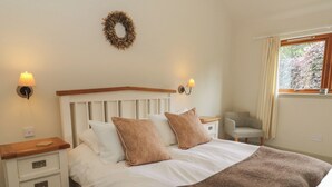Une chambre à coucher avec une tête de lit en bois, un lit blanc, deux oreillers, un plaid marron, une table de chevet avec des tiroirs, une chaise, une fenêtre avec des rideaux et un luminaire fixé au mur.