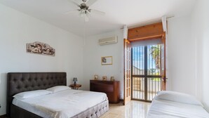 Quarto com uma cama grande, uma cama menor, uma mesinha de cabeceira, cômoda e um ventilador de teto.