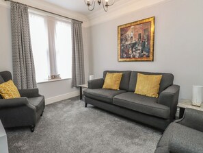 A living room with a grey sofa, patterned curtains, a painting, and a small table with a lamp. - Conway Mart Cottage (Conwy)