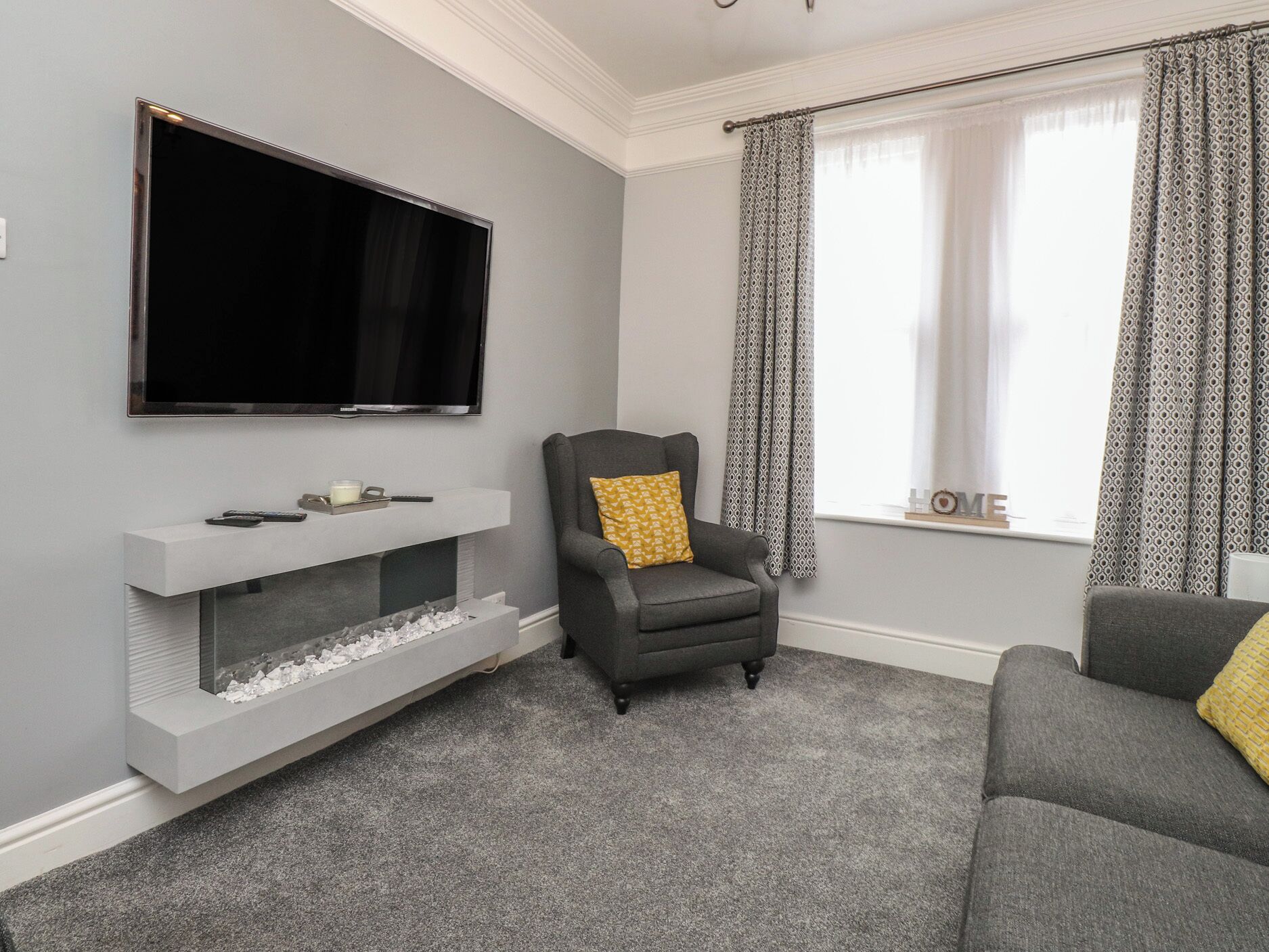 A living room with a flat-screen TV mounted on the wall, a grey armchair with a yellow pillow, a grey sofa, and a white entertainment unit with a fireplace.