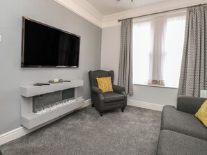 Een woonkamer met een flatscreen televisie aan de muur, een grijze fauteuil met een geel kussen, een grijze bank en een witte tv-kast met een open haard.