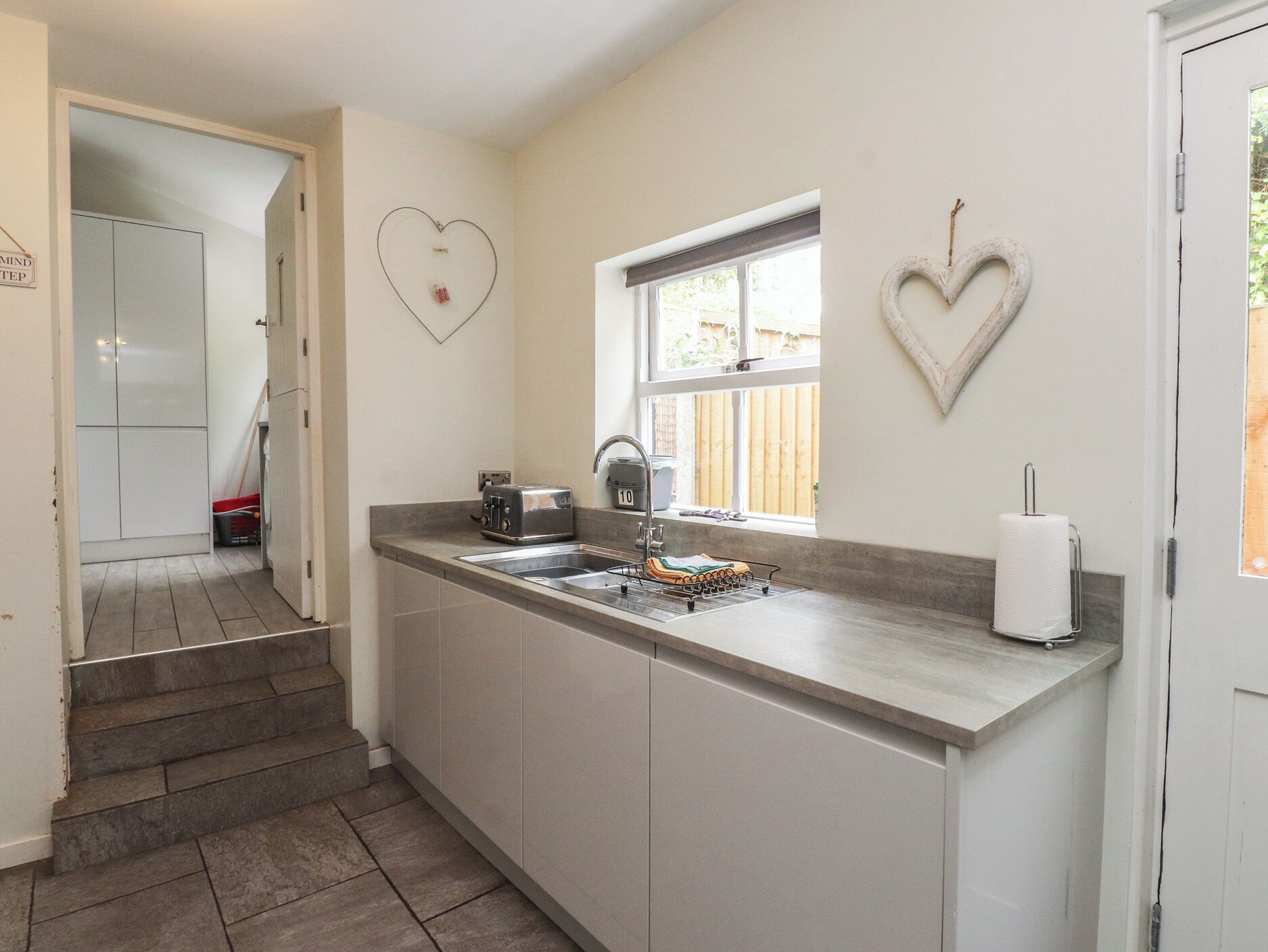A kitchen with a double sink, a toaster, a window, and a heart-shaped decoration on the wall.