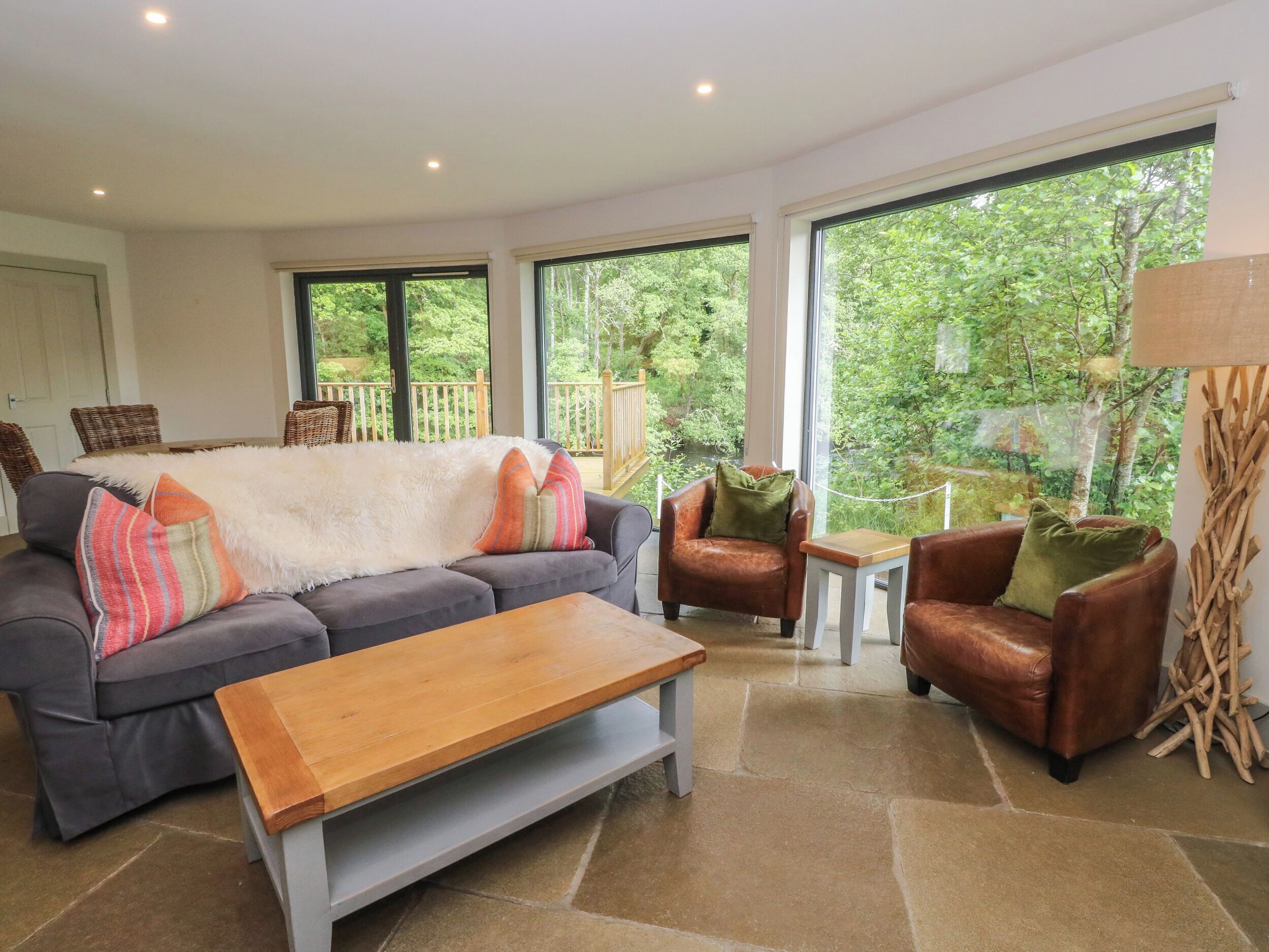 Sala de estar con un sofá gris, una mesa de centro de madera, un sillón de cuero marrón y una pequeña mesa blanca. Amplias ventanas ofrecen vistas al verde exterior.