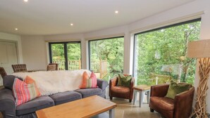 A living room with a grey sofa, a wooden coffee table, a brown leather armchair, and a small white table. Large windows provide a view of greenery outside. - The Crannog (Killin)