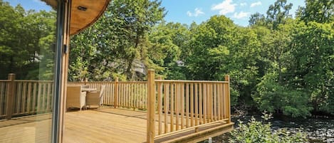 A wooden deck with a glass enclosure, surrounded by lush greenery and a river.