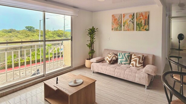 A modern living room with a large window, a sofa with patterned cushions, a wooden coffee table, and a potted plant. - Modern Apartment with Pool Near Unicentro Shopping Center (Neiva)