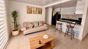 A modern living room with a sofa, a coffee table, a potted plant, and a kitchen area with a refrigerator and bar stools. - Modern Apartment with Pool Near Unicentro Shopping Center (Neiva)