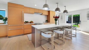 A modern kitchen with wooden cabinets, a white countertop, stainless steel bar stools, pendant lights, and a large window. - The Orient Retreat - Luxury Designer Retreat with Water Views in Orient, NY (Orient)