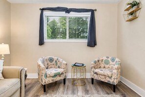 A room with two patterned armchairs, a small round table, a window with blinds, and a wall-mounted plant shelf.