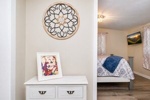 Une chambre à coucher avec un lit, une table de chevet sur laquelle est posée une photo encadrée, un  élément décoratif mural et un tableau accroché au mur.