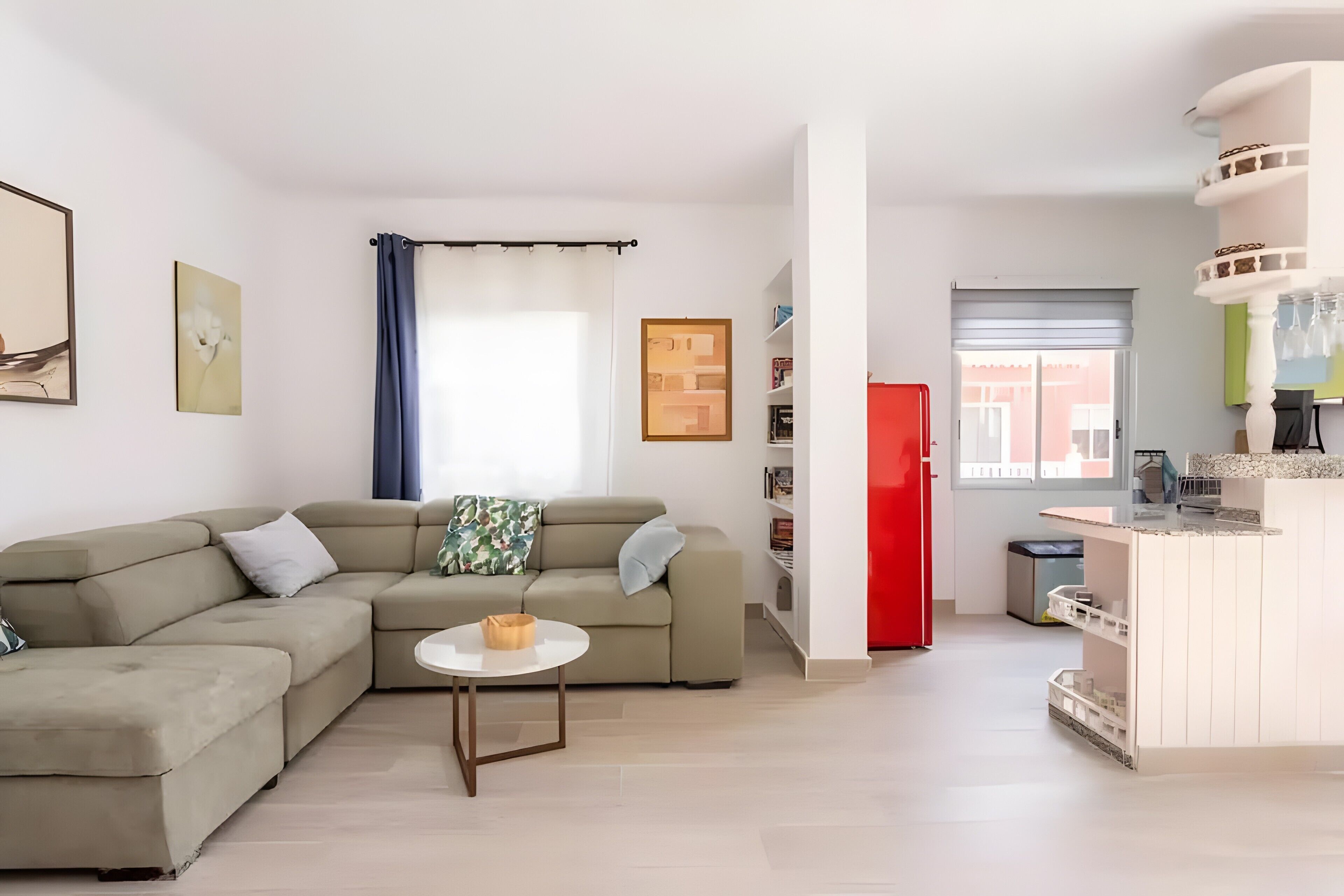 Een moderne woonkamer met een hoekbank, een klein rond tafeltje en een keukenhoek met een rode koelkast.