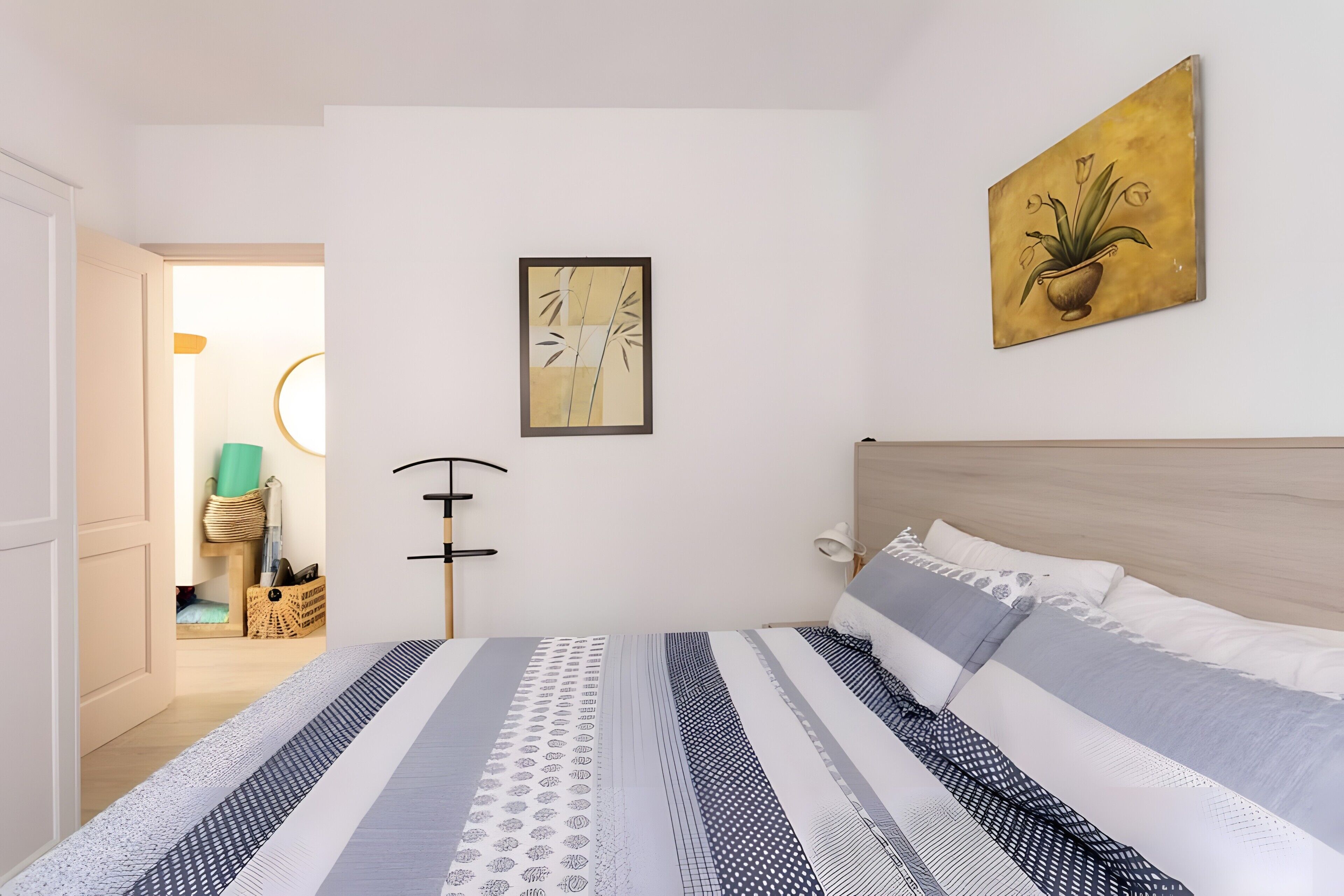 Een slaapkamer met een bed, nachtkastje, lamp, een schilderij aan de muur en een deur naar een andere kamer.