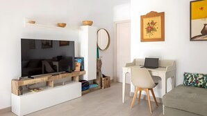 Un salon moderne avec un téléviseur à écran plat, un bureau blanc avec un ordinateur portable, un canapé beige et une petite table sur laquelle se trouve un bol.