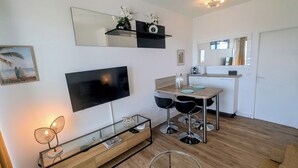 Een moderne woonkamer met een flatscreen televisie, een eethoek met tafel en stoelen, een keuken met aanrecht en kasten, en een grote spiegel aan de muur.