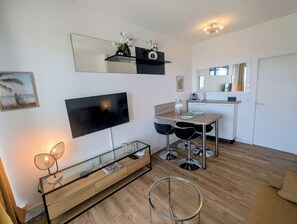 Ein modernes Wohnzimmer mit Flachbildfernseher, Essbereich mit Tisch und Stühlen, Küchenzeile mit Arbeitsplatte und Schränken sowie ein großer Spiegel an der Wand.