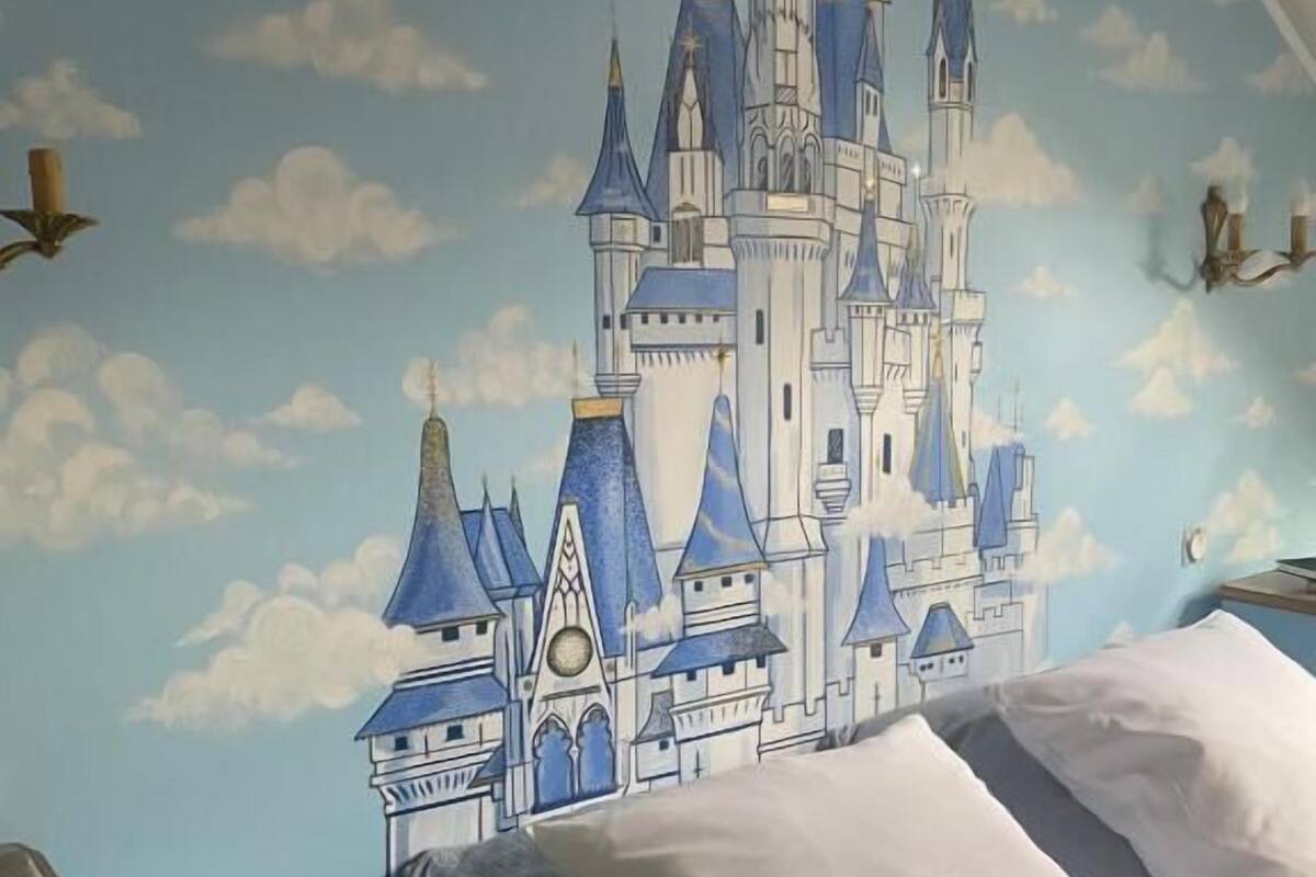 Un dormitorio con un mural de castillo en la pared, una cama con ropa blanca y una mesita de noche con lámpara.