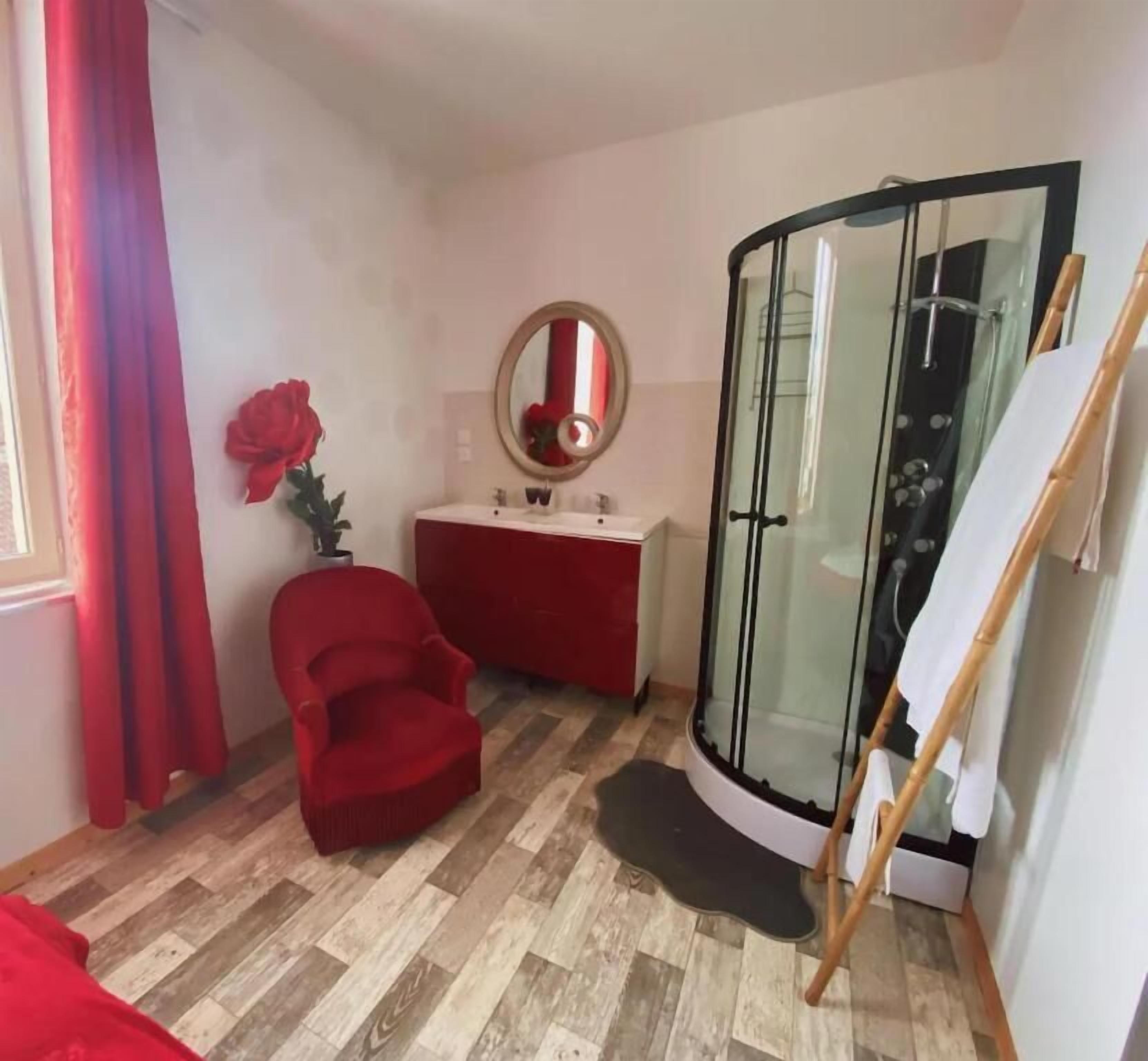 Un baño con mampara de vidrio, una silla roja, un tocador con espejo y una decoración floral roja.