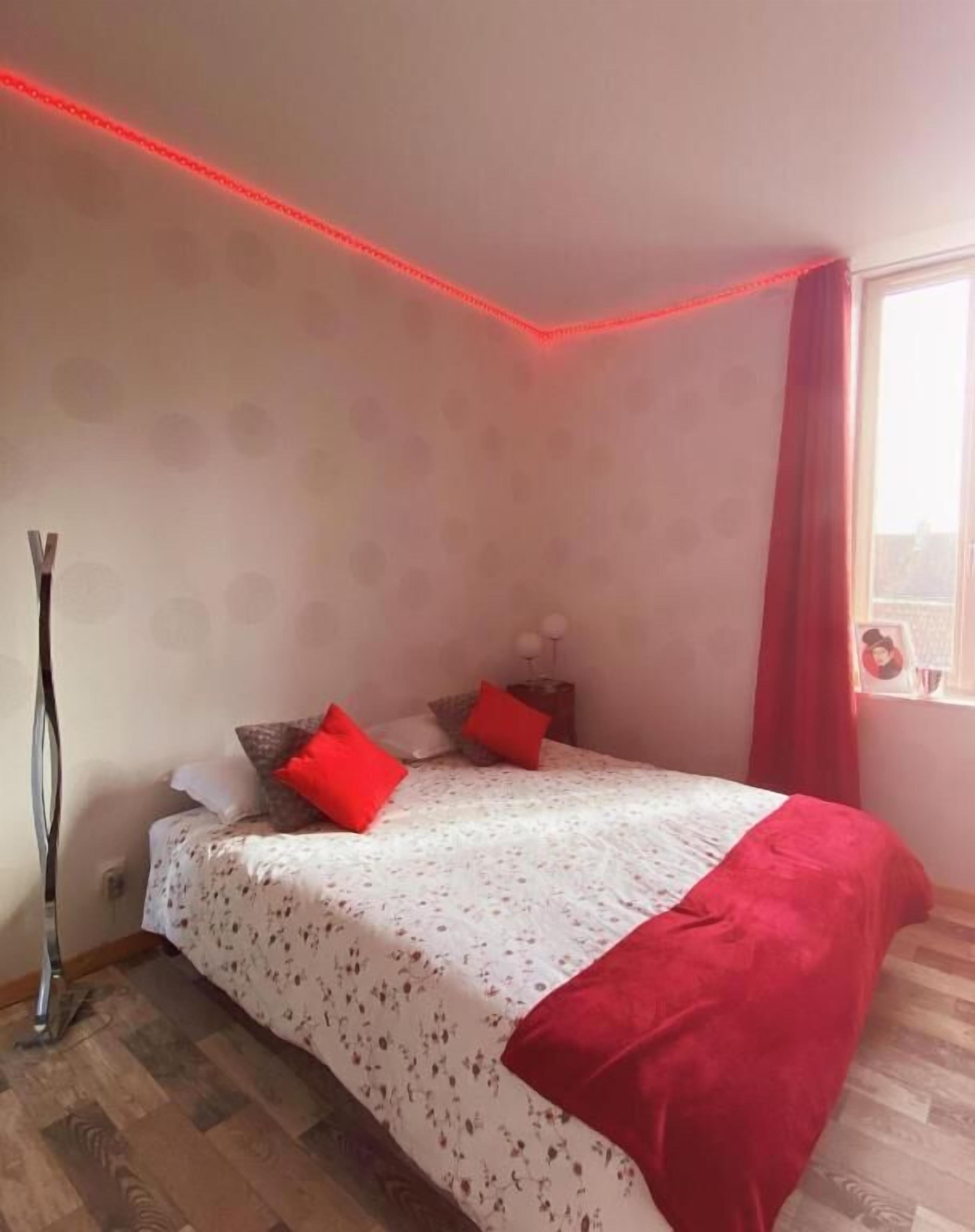 Un dormitorio con cama, mesita de noche, lámpara, ventana con cortinas y una tira de luces LED rojas a lo largo del techo.