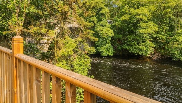 A wooden bridge over a river with dense green foliage on both sides. - Otters Holt (Killin)
