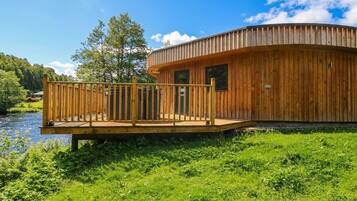 A wooden cabin with a deck, a river, and trees in the background.