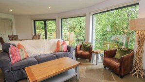 A living room with a sofa, two armchairs, a coffee table, and a view of greenery through large windows. - Otters Holt (Killin)