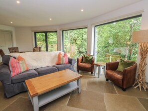 A living room with a sofa, two armchairs, a coffee table, and a view of greenery through large windows.