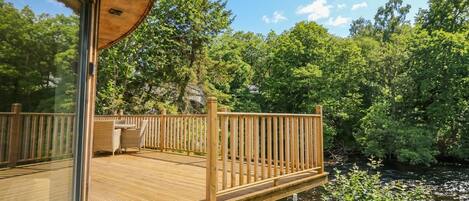 A wooden deck with a glass enclosure, surrounded by lush greenery and a river.