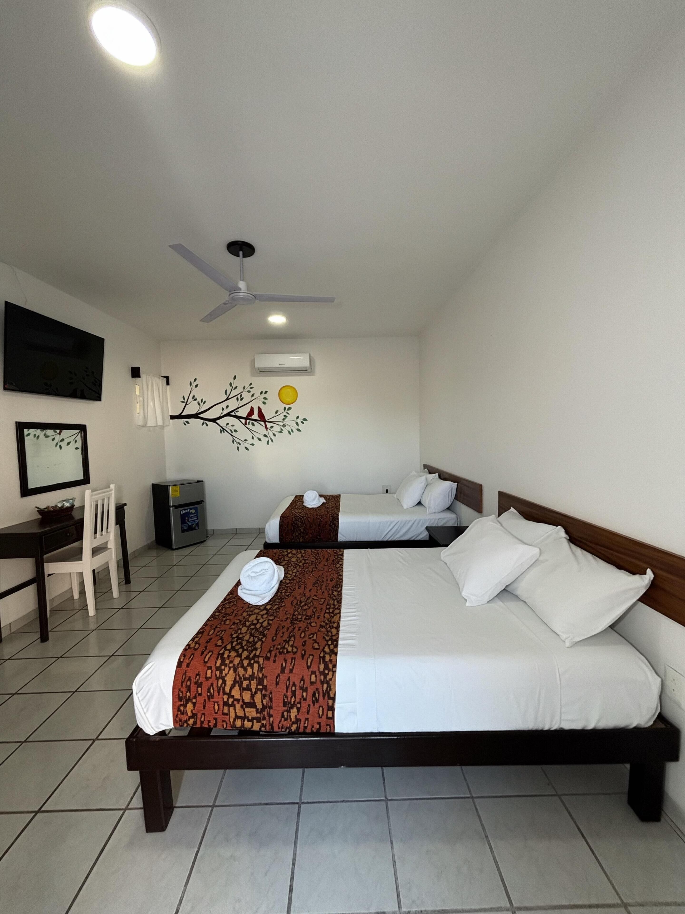 Habitación de hotel con dos camas, un escritorio, una silla, ventilador de techo y aire acondicionado instalado en la pared.