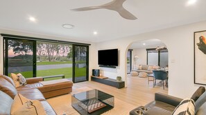 A modern living room with a large TV, comfortable seating, and a view of a green landscape. - Salthill - Waterfront Pet Friendly Family Home on the Tweed River - Min 3N (Tweed Heads)