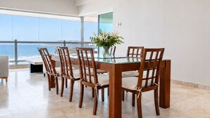 A dining area with a glass table and wooden chairs, a vase with flowers, and a view of the ocean.