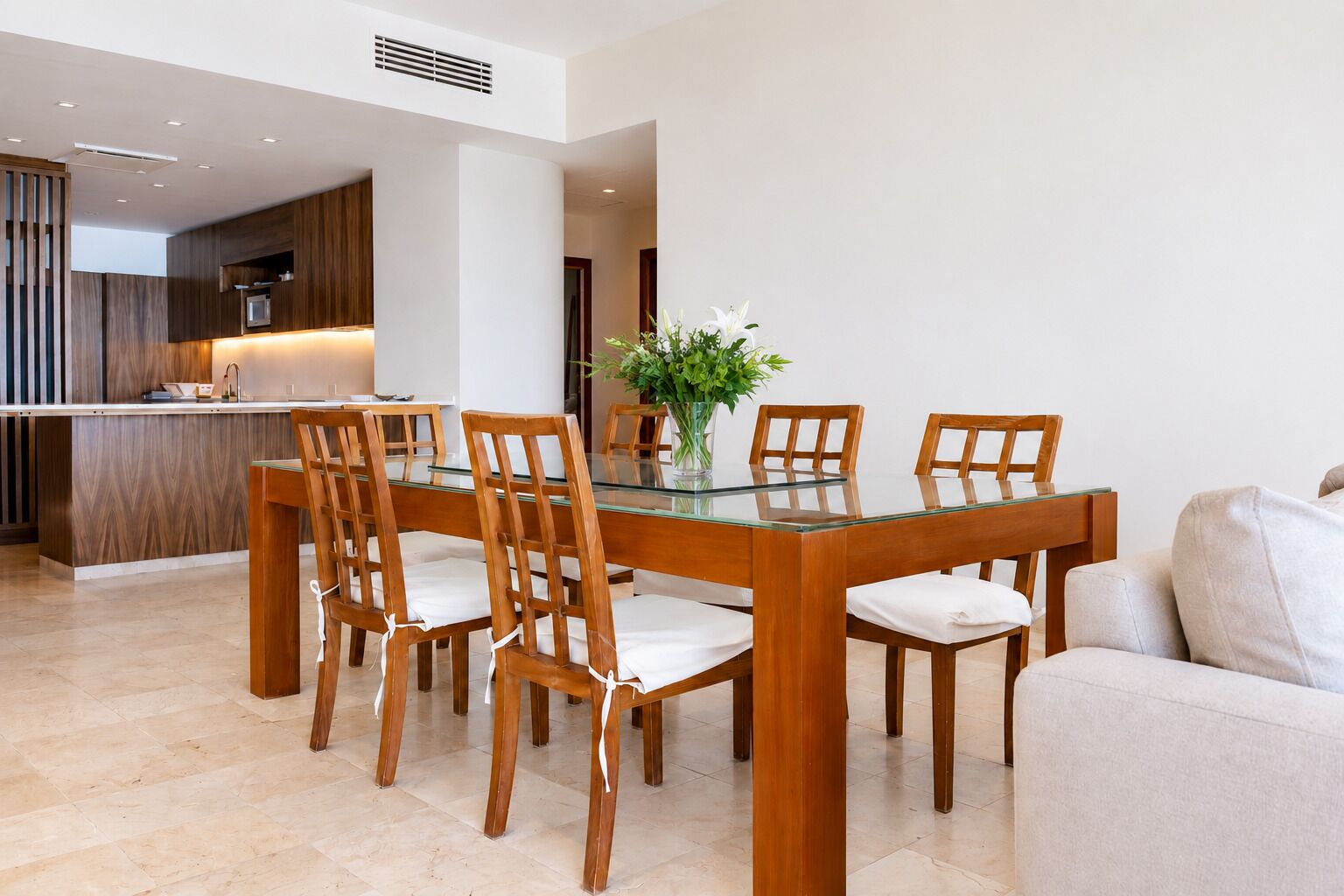Uma área de jantar moderna com mesa de vidro e cadeiras de madeira, cozinha ao fundo e um sofá em primeiro plano.