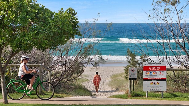 Une plage avec un panneau indiquant l’absence de drapeaux et un avertissement concernant la baignade, une personne qui se promène et un cycliste.