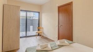 Quarto com cama, guarda-roupa de madeira, porta e uma cadeira perto de uma porta de vidro deslizante que dá para o exterior.